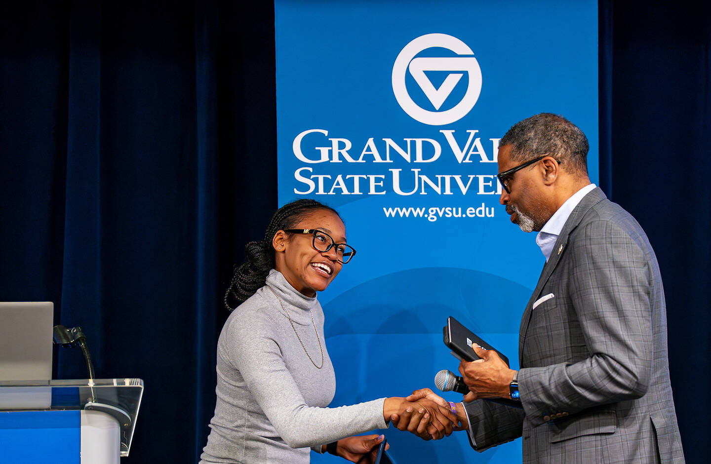 A Student in a gray sweater shakes hands with another person in a suit at a Grand Valley State University event, with the university's logo and website visible on a blue backdrop.